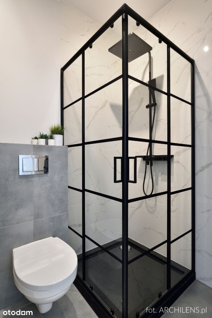 Nowoczesna łazienka z kabiną prysznicową typu walk-in o czarnych, geometrycznych ramach, marmurowe płytki na ścianach, podwieszana toaleta i trzy małe rośliny doniczkowe na półce.