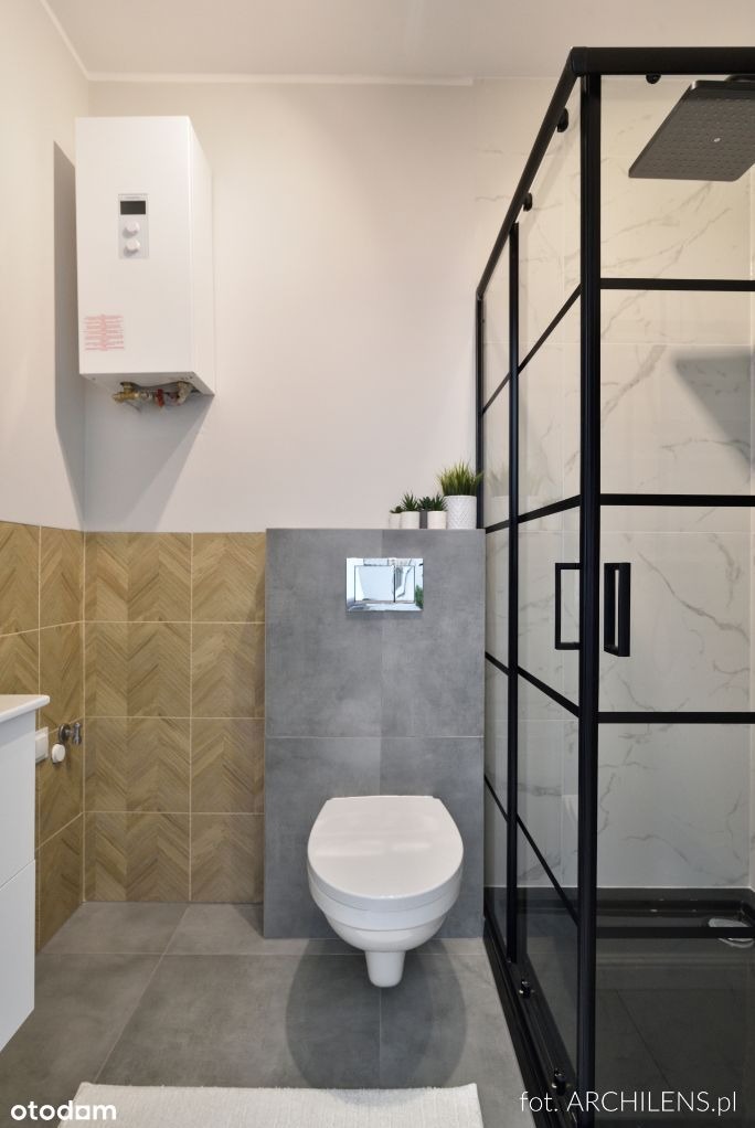 Nowoczesna łazienka z kabiną prysznicową typu walk-in z czarnymi ramami, wiszącą toaletą, płytkami imitującymi drewno w jodełkę i grzejnikiem nad zabudową.