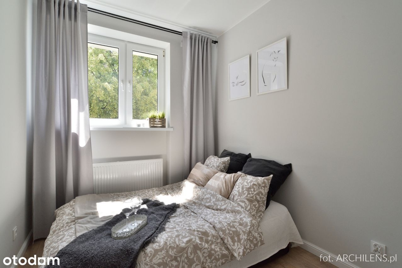 Jasny pokój z łóżkiem, szarymi zasłonami i dekoracyjnymi obrazkami na ścianie, widok na zieleń za oknem.