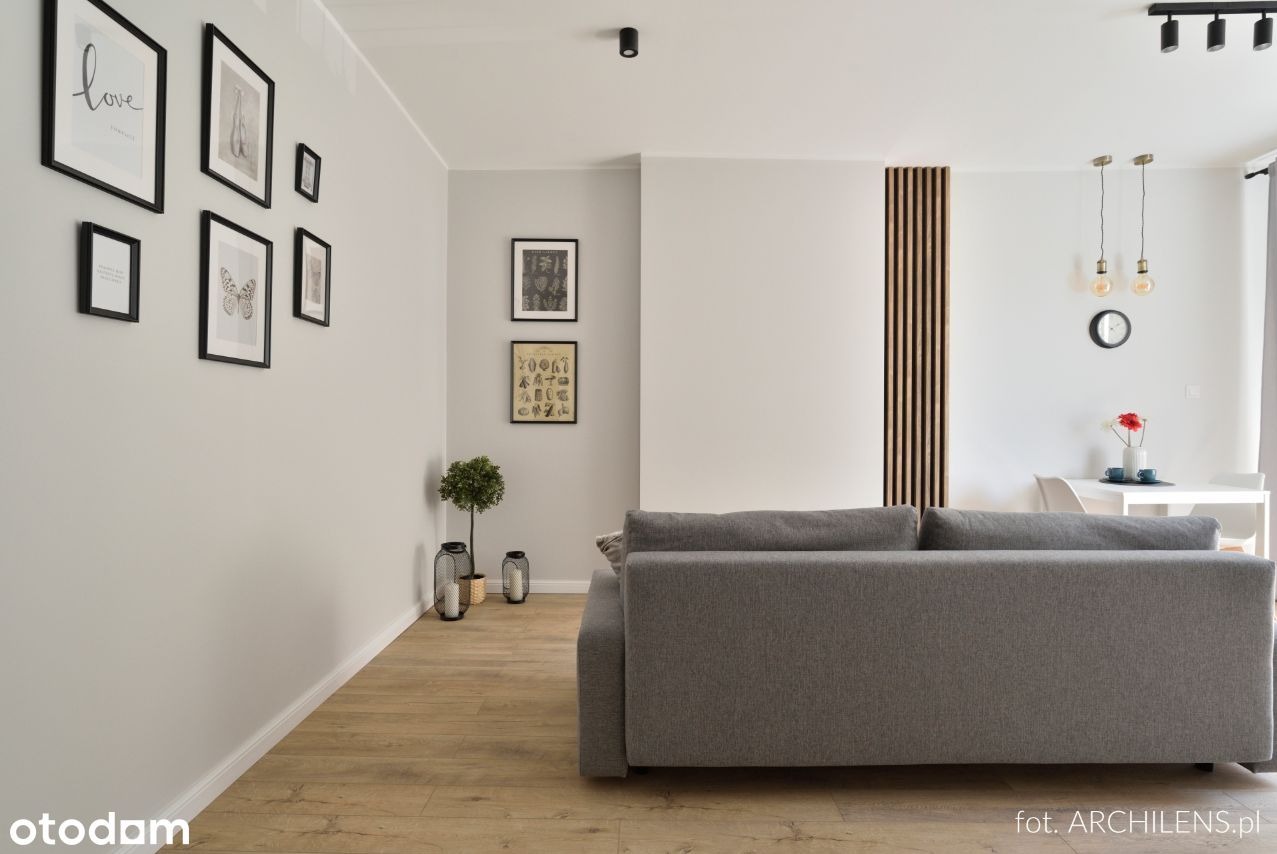 Jasne wnętrze salonu z szarą sofą, drewnianą podłogą i galerią czarno-białych obrazków na ścianie, uzupełnione dekoracyjnymi lampionami i rośliną w doniczce.