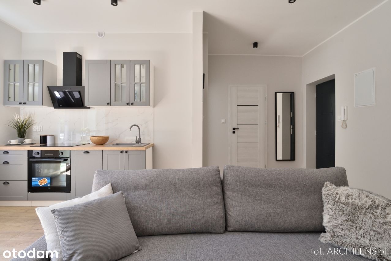 Nowoczesna kuchnia w odcieniach szarości z drewnianymi blatami, widok z salonu na zabudowę kuchenną z piekarnikiem, płytą indukcyjną i okapem, w tle biała ściana z drzwiami.