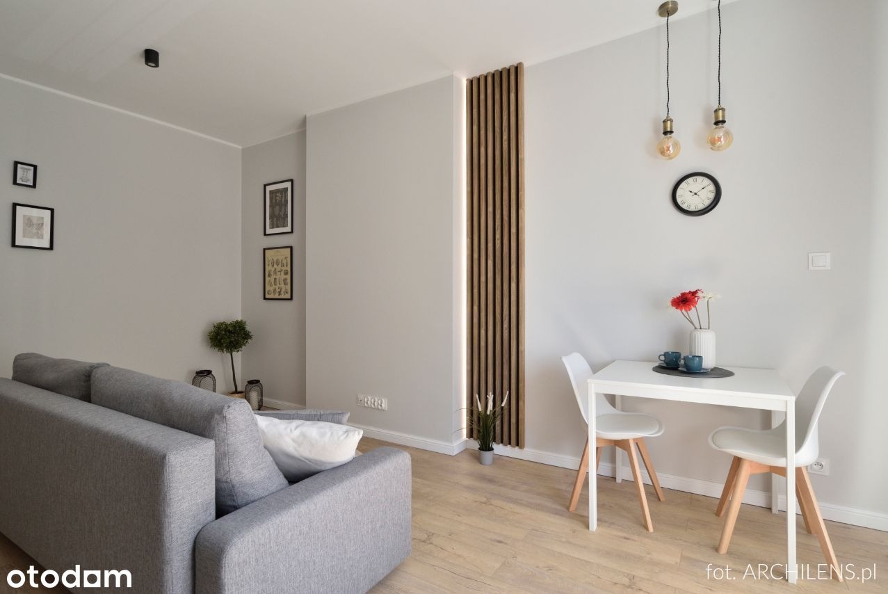 Nowoczesny salon z szarą sofą, drewnianymi lamelami na ścianie z podświetleniem LED, białym stołem z krzesłami i dekoracjami, oświetlony wiszącymi lampami z żarówkami Edisona.