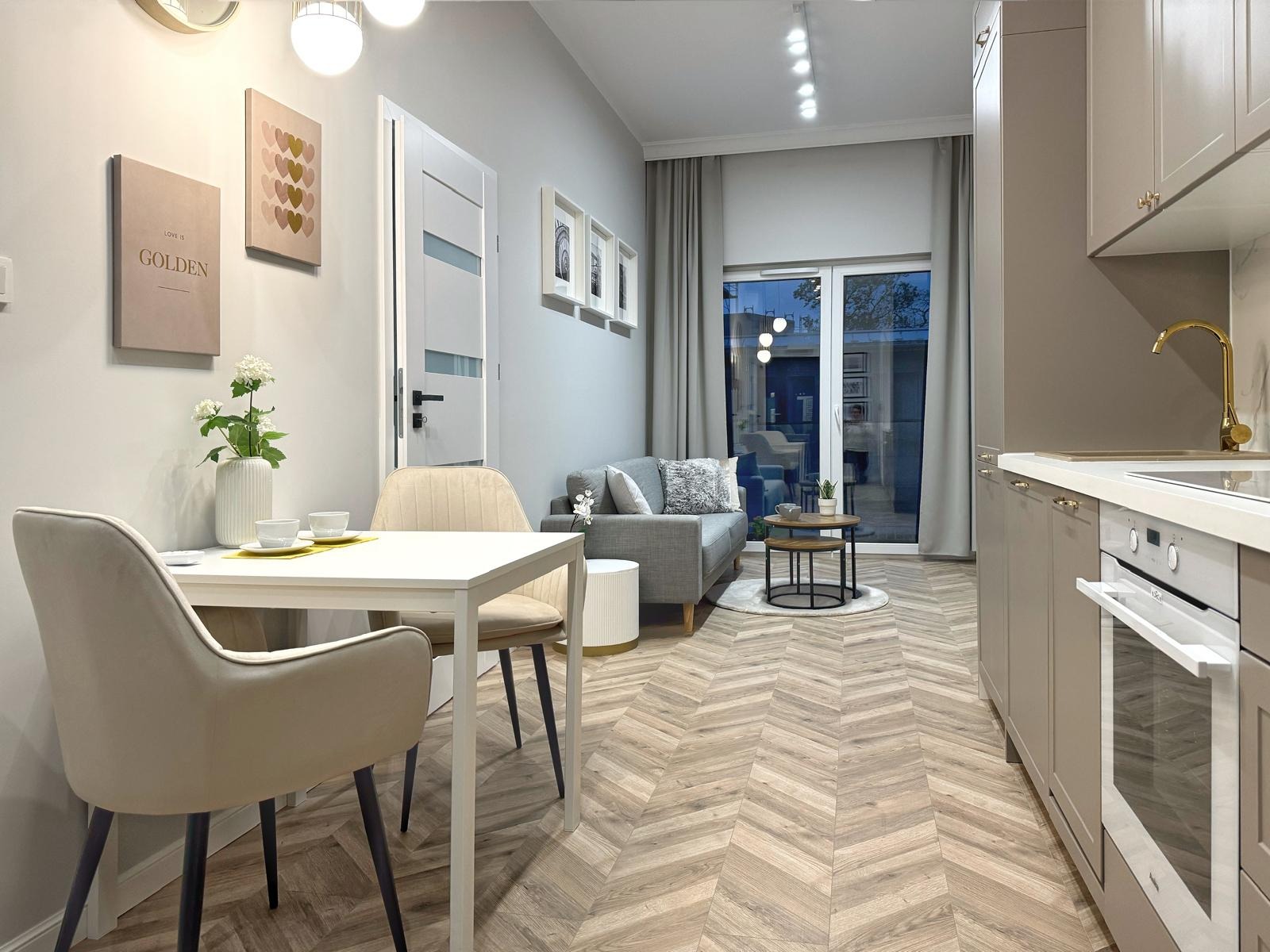 Jasne wnętrze mieszkania po remoncie: parkiet w jodełkę, stół z kwiatami, szara sofa i zabudowa kuchenna z baterią w kolorze złotym.