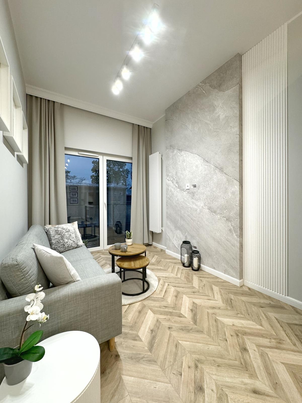 Salon w odcieniach szarości i beżu z parkietem ułożonym w jodełkę, szarą sofą, okrągłymi stolikami kawowymi i ścianą z imitacji marmuru.