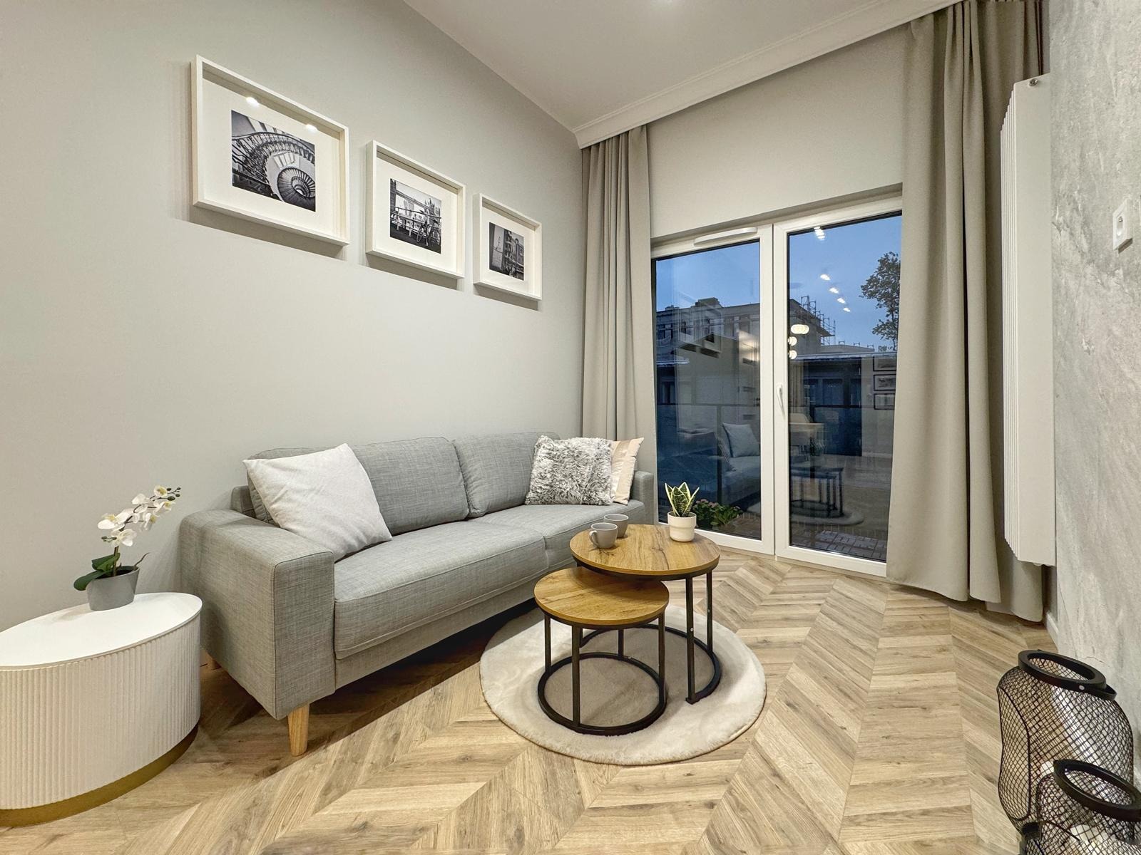 Jasne wnętrze salonu z szarą sofą, drewnianą podłogą w jodełkę, okrągłymi stolikami kawowymi i trzema czarno-białymi zdjęciami w białych ramach na ścianie.