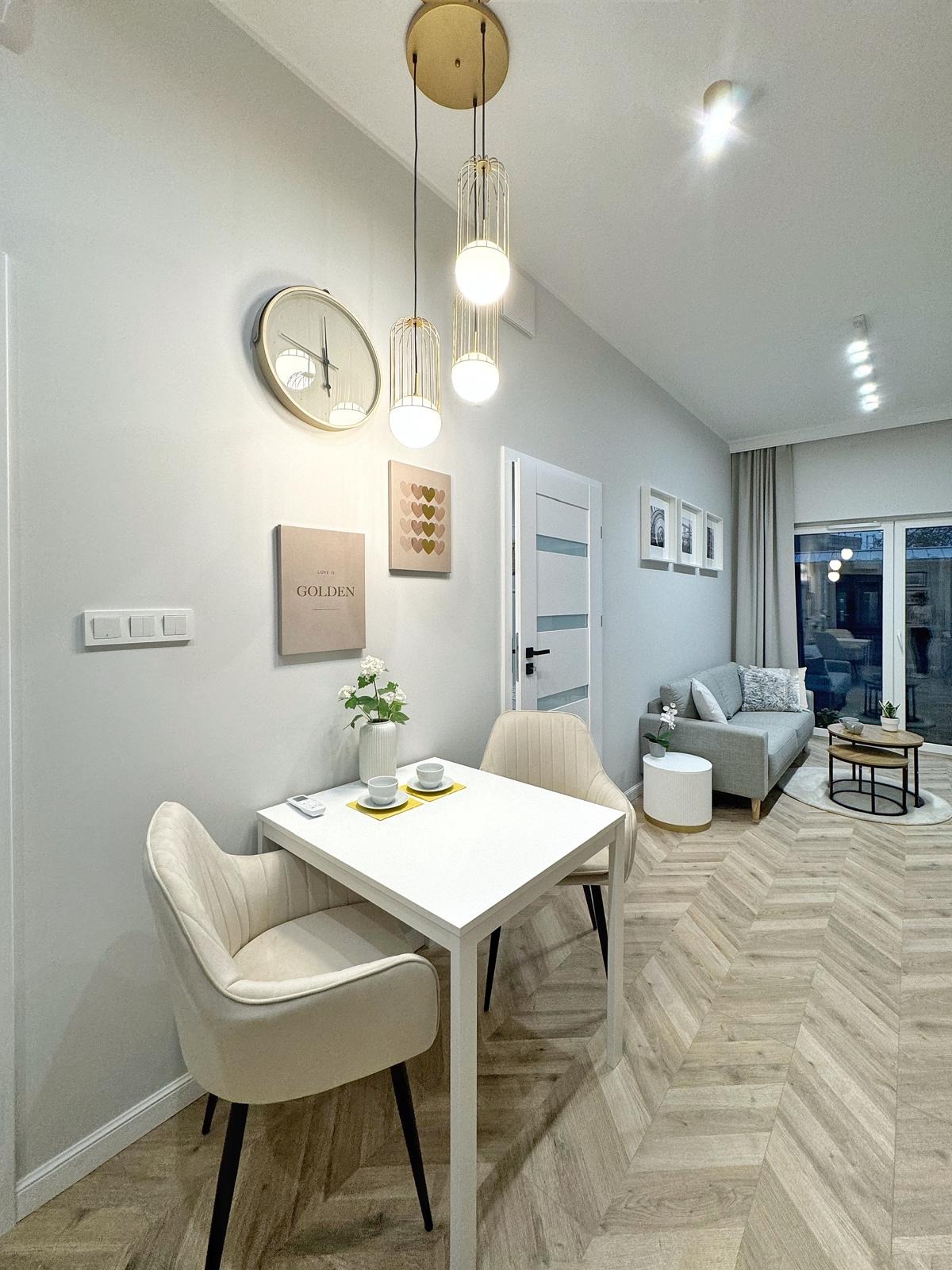 Jasne wnętrze mieszkania: stół z dwoma krzesłami, dekoracje ścienne, złoty zegar, designerskie lampy wiszące i parkiet ułożony w jodełkę.