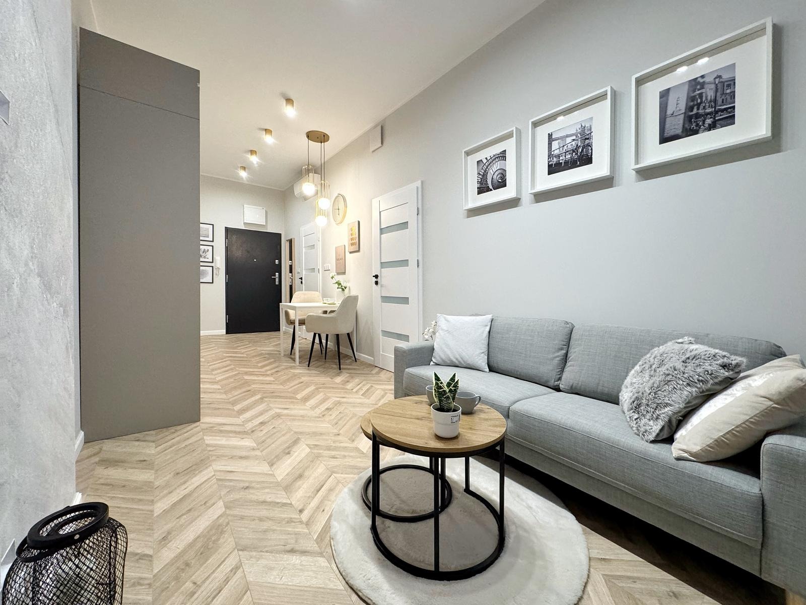 Jasne wnętrze mieszkania z parkietem w jodełkę, szarą sofą, okrągłym stolikiem kawowym i trzema czarno-białymi zdjęciami w białych ramkach na ścianie.