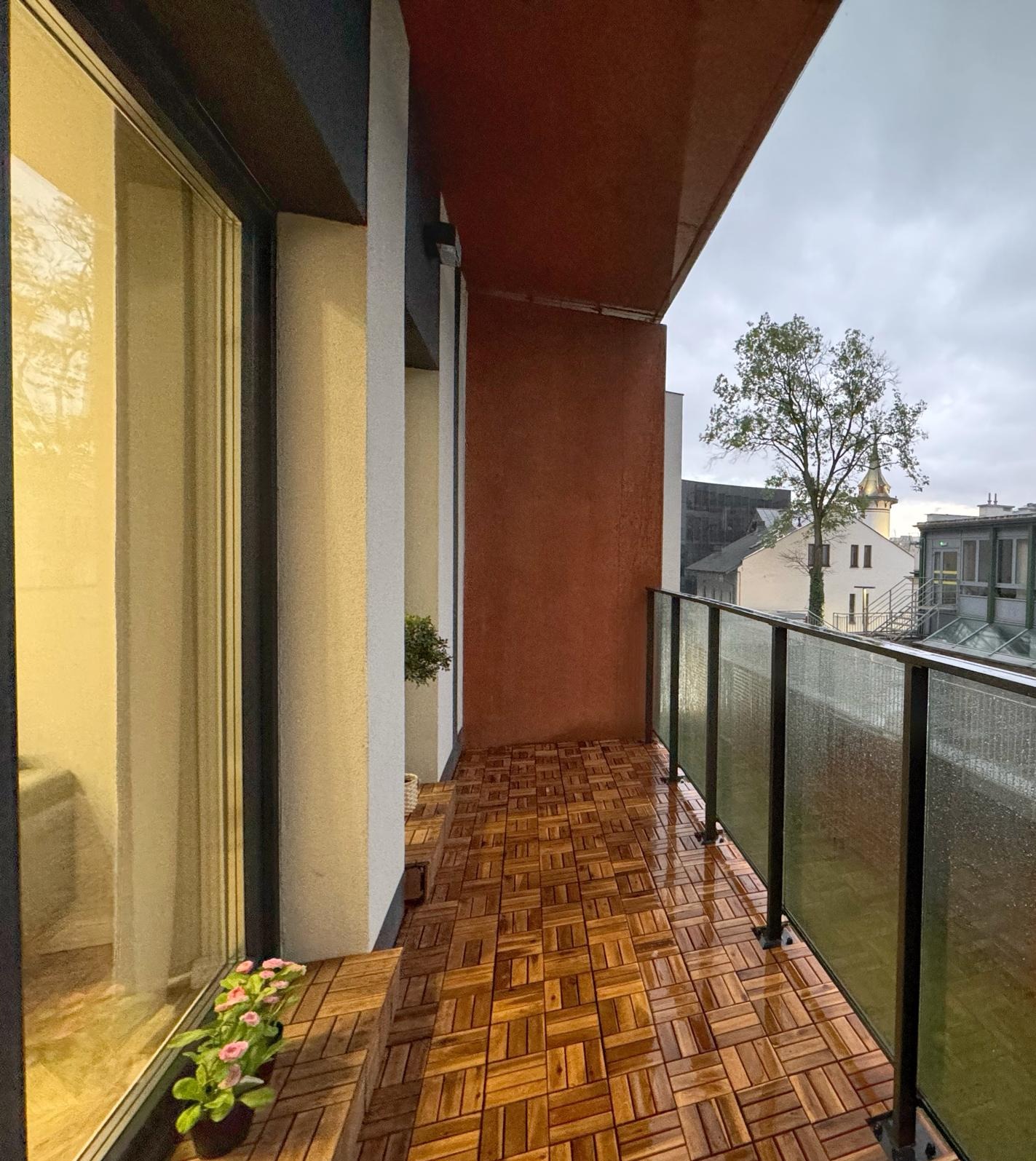 Balkon wyłożony drewnianą kostką, z widokiem na miasto w pochmurny dzień, szklana balustrada z kroplami deszczu, doniczka z różowymi kwiatami przy oknie.