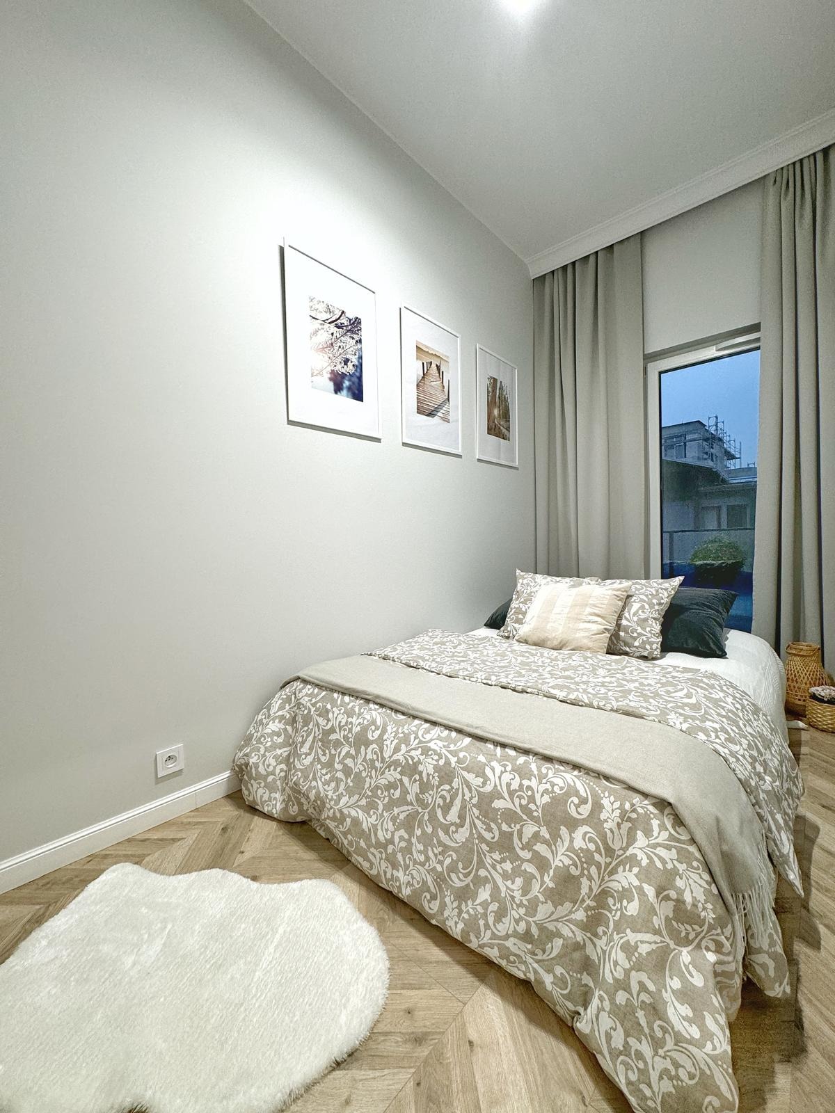 Jasna sypialnia z łóżkiem w beżowej pościeli z ornamentami, drewnianą podłogą w jodełkę, białym dywanikiem w kształcie chmurki i trzema obrazami w białych ramkach na szarej ścianie.