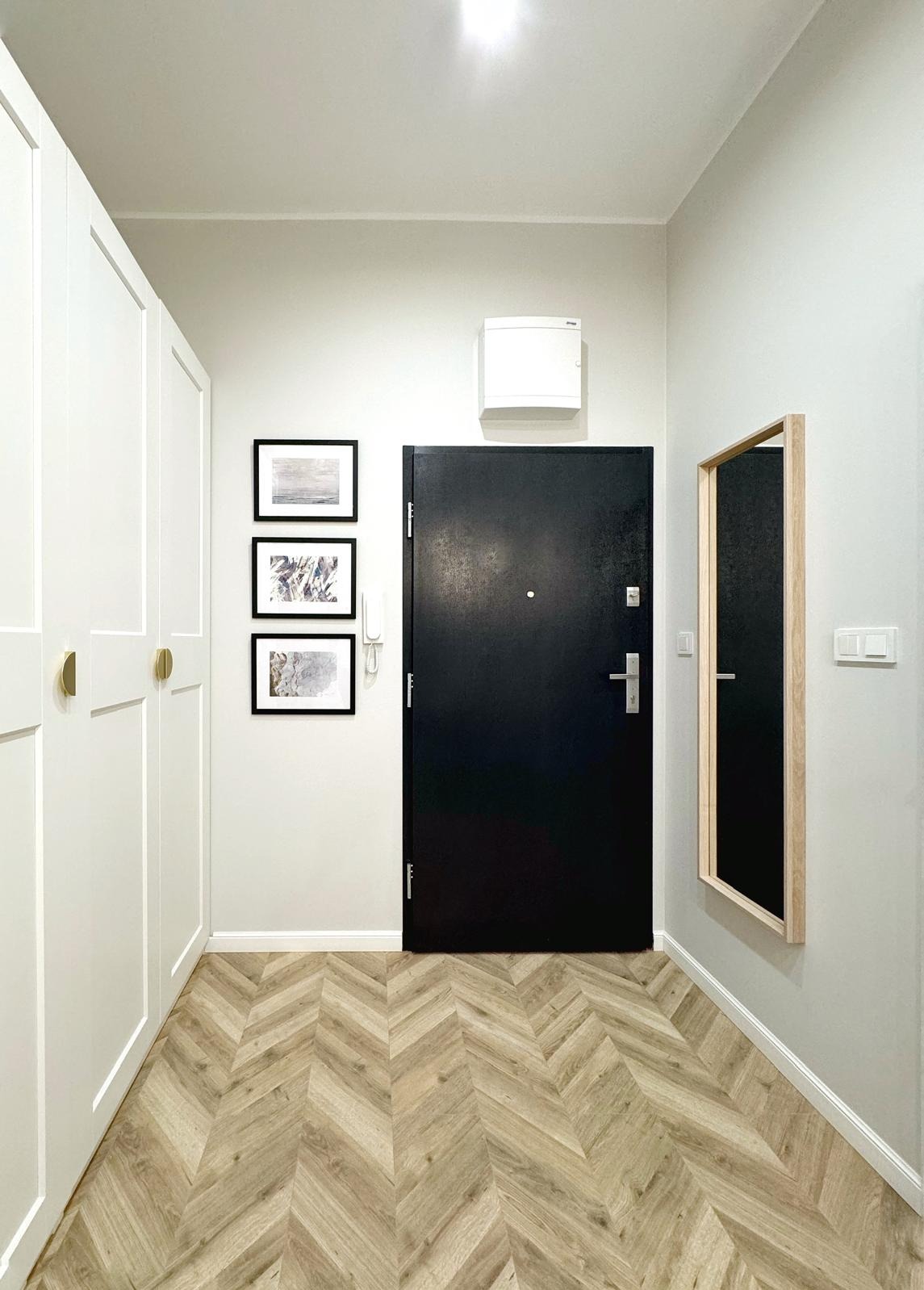 Nowoczesny przedpokój z czarnymi drzwiami wejściowymi, podłogą w jodełkę, białymi szafami w zabudowie i lustrem w drewnianej ramie.
