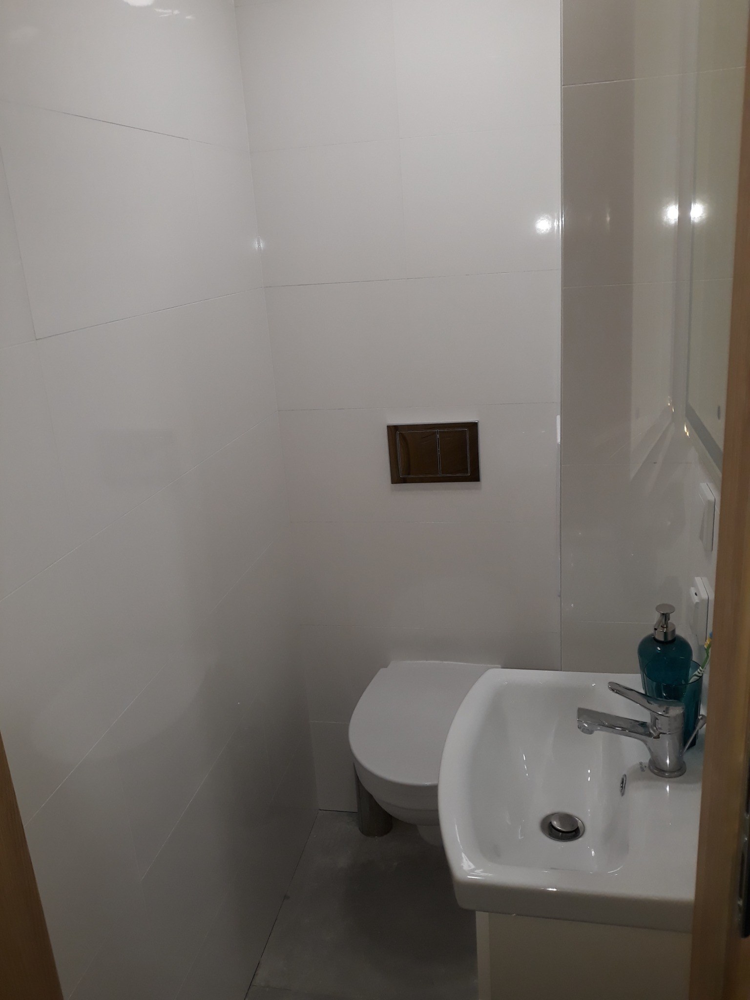 Biała, mała łazienka wykończona płytkami ceramicznymi, z wiszącą miską WC i umywalką z baterią oraz dozownikiem mydła w kolorze morskim.