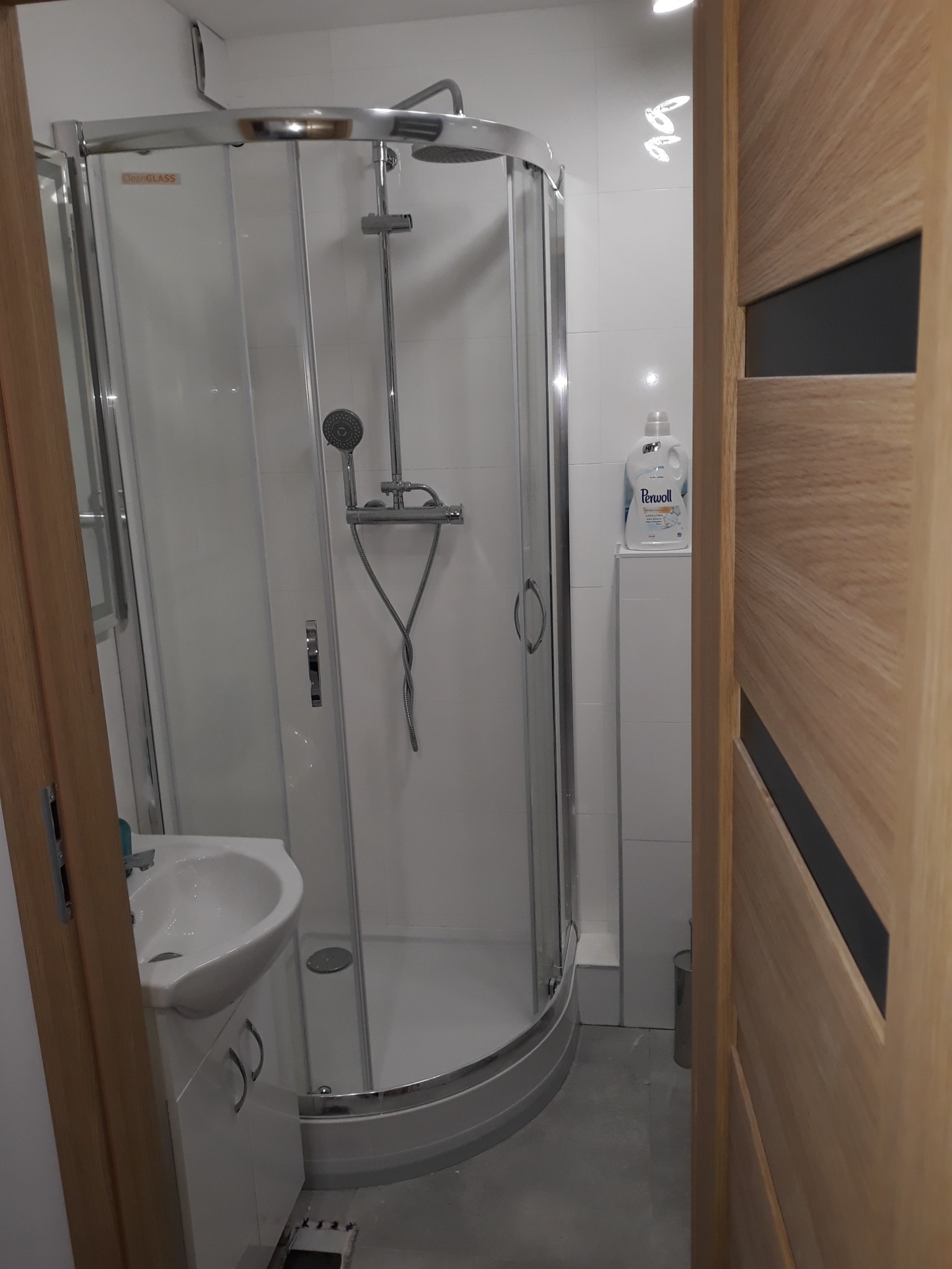 Nowoczesna kabina prysznicowa z chromowanymi elementami, biała umywalka z szafką, widoczna butelka płynu do prania Perwoll, drzwi z drewnianą okleiną i czarnymi wstawkami, szare płytki podłogowe.