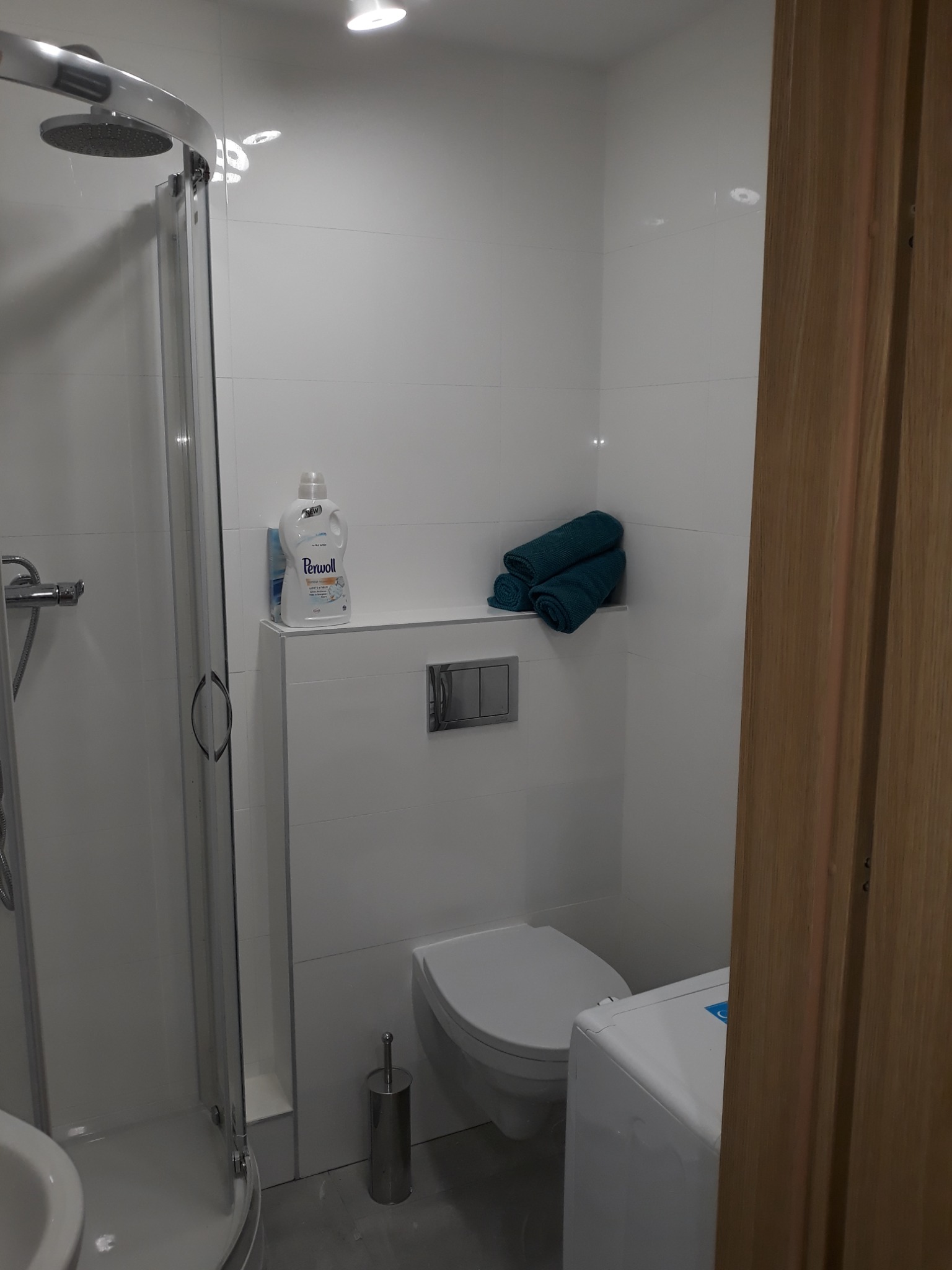 Nowoczesna, schludna łazienka z kabiną prysznicową, podwieszaną toaletą, pralką i stertą turkusowych ręczników na półce, uzupełniona butelką płynu do prania Perwoll.