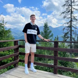 Młody mężczyzna w koszulce Yale University stoi na drewnianym tarasie z widokiem na góry i las, błękitne niebo z chmurami w tle.