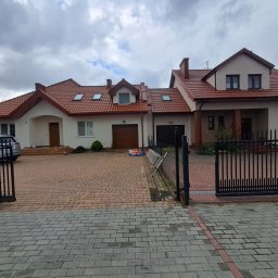 Kacper - Dwukondygnacyjny dom z czerwoną dachówką, widoczny zza otwartej, czarnej bramy. Na podjeździe drabina i puszki z farbą. Pochmurne niebo w tle.