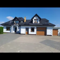 Biały dom jednorodzinny z szarymi akcentami, czarnym dachem i panelami słonecznymi, sfotografowany w słoneczny dzień. Widoczne drewniane okna i drzwi garażowe.