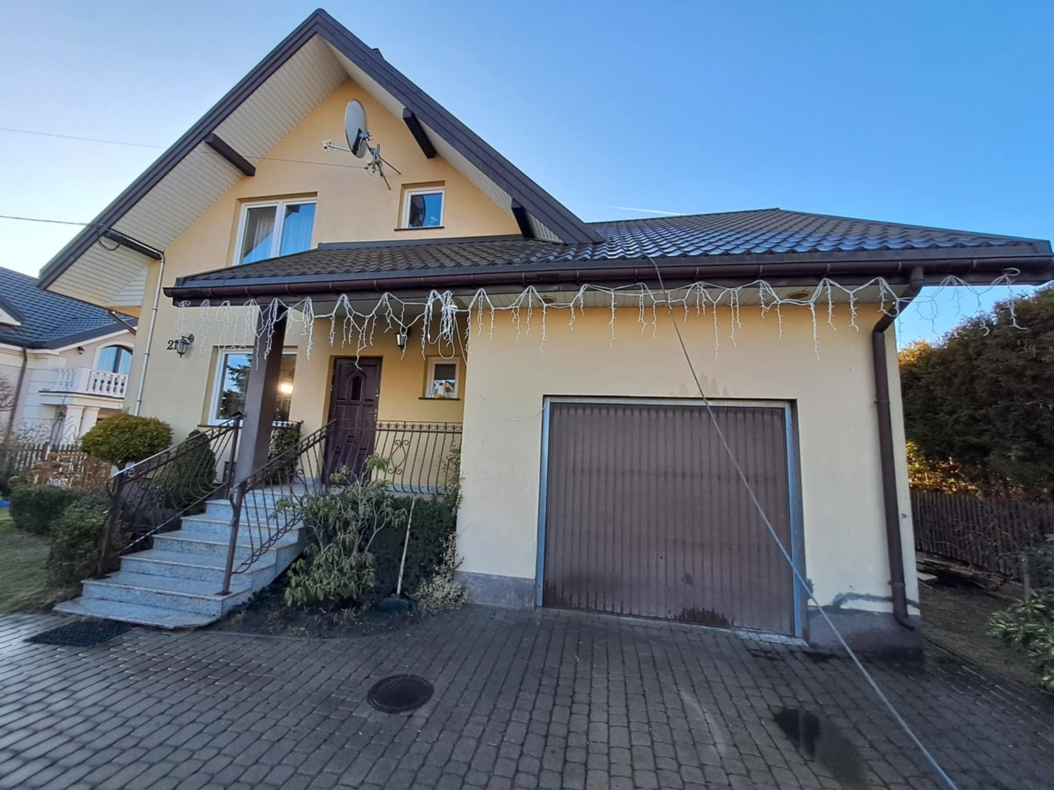 Dwukondygnacyjny dom z beżową elewacją, brązowym dachem i garażem. Schody wejściowe z metalową balustradą. Dekoracyjne lampki zwisają z dachu. Widok z poziomu brukowanego podjazdu.