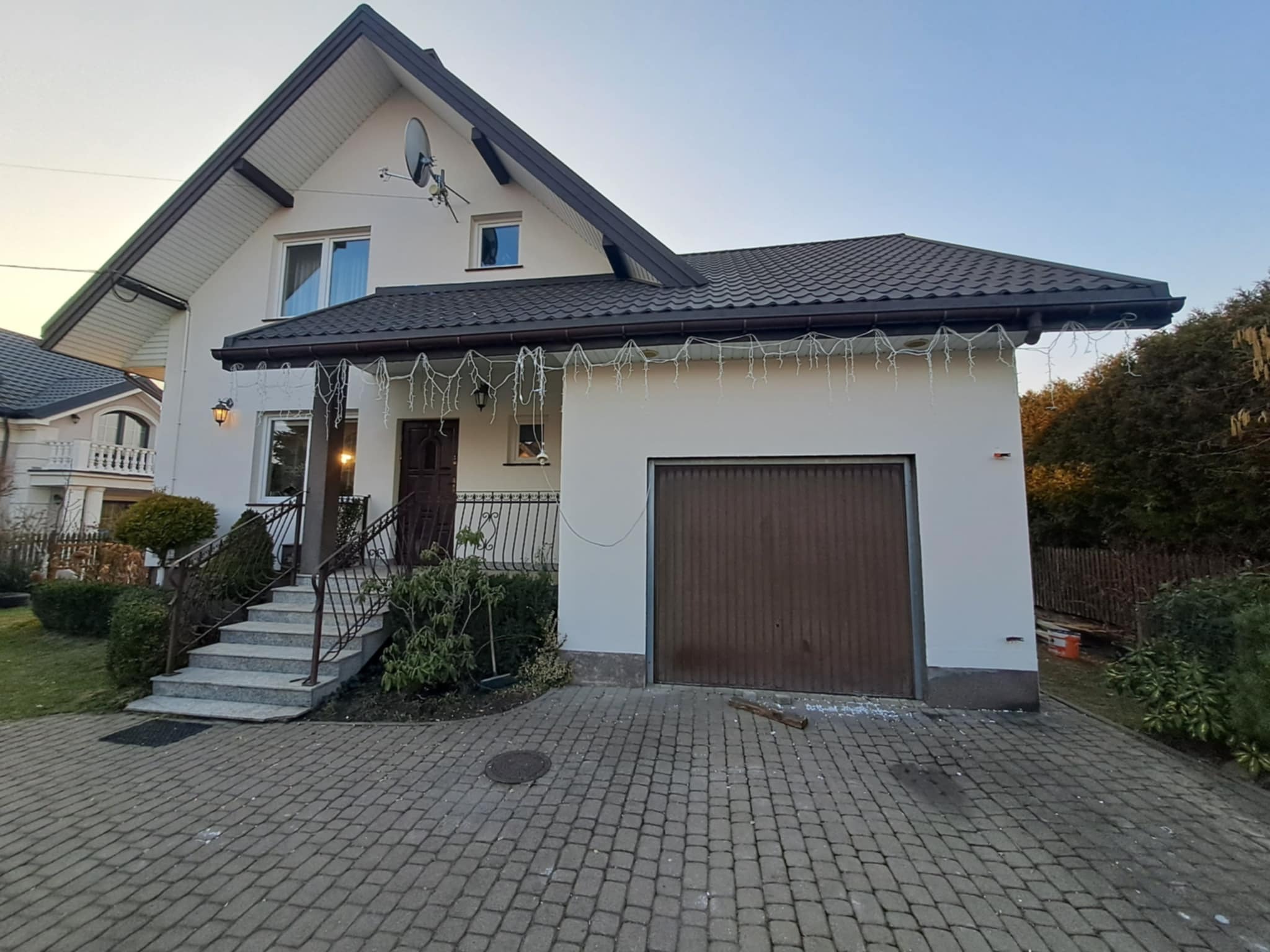 Jasny dom jednorodzinny z brązowym dachem i garażem, oświetlony wieczornym światłem. Widoczne schody wejściowe i podjazd z kostki brukowej.