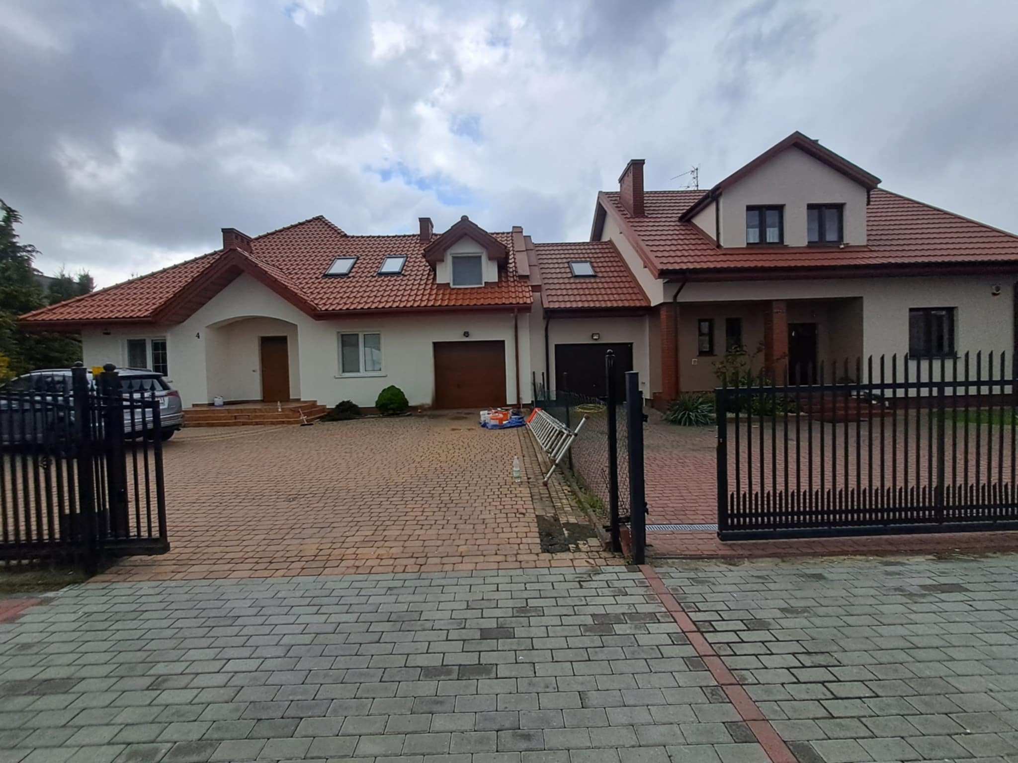 Dwukondygnacyjny dom z czerwoną dachówką, widoczny zza otwartej, czarnej bramy. Na podjeździe drabina i puszki z farbą. Pochmurne niebo w tle.