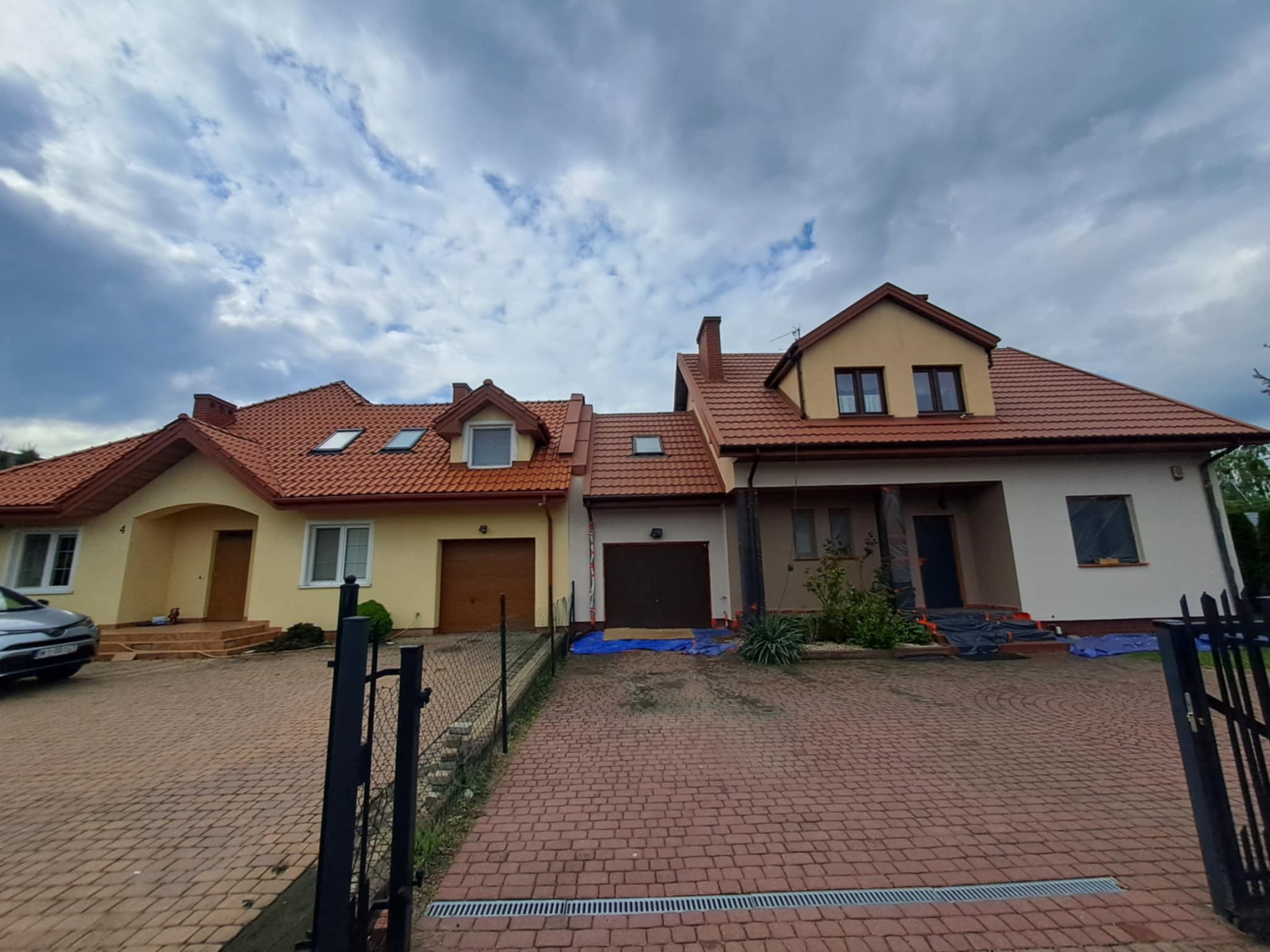 Dwa domy jednorodzinne z czerwonym dachem, jeden w trakcie przygotowania do malowania elewacji, zabezpieczony folią, widoczne elementy rusztowania.