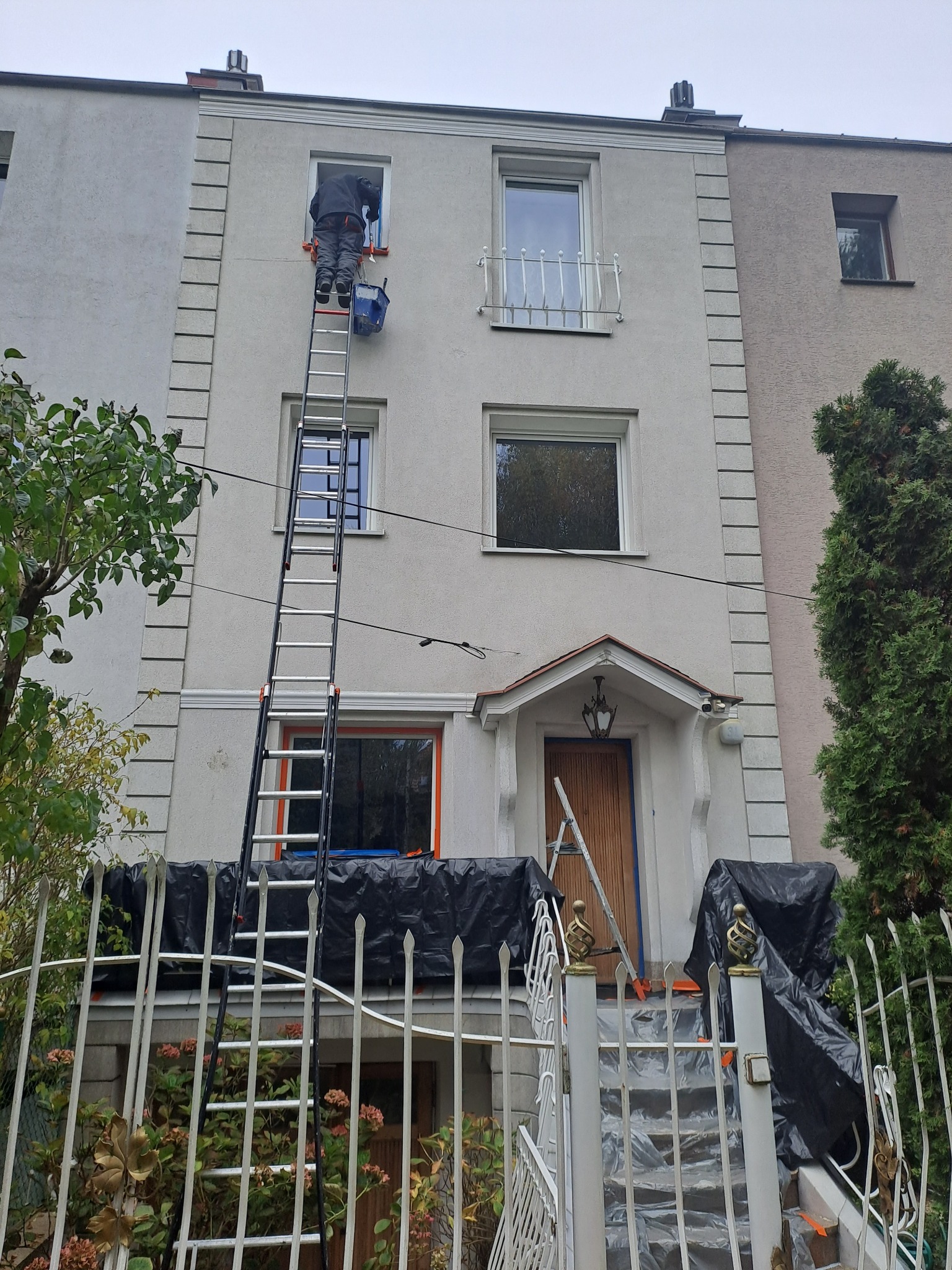 Pracownik na drabinie maluje elewację trzypiętrowego domu z dekoracyjnymi elementami. Widoczne zabezpieczenia i narzędzia malarskie. Dom sąsiaduje z budynkami o różnej strukturze fasady.
