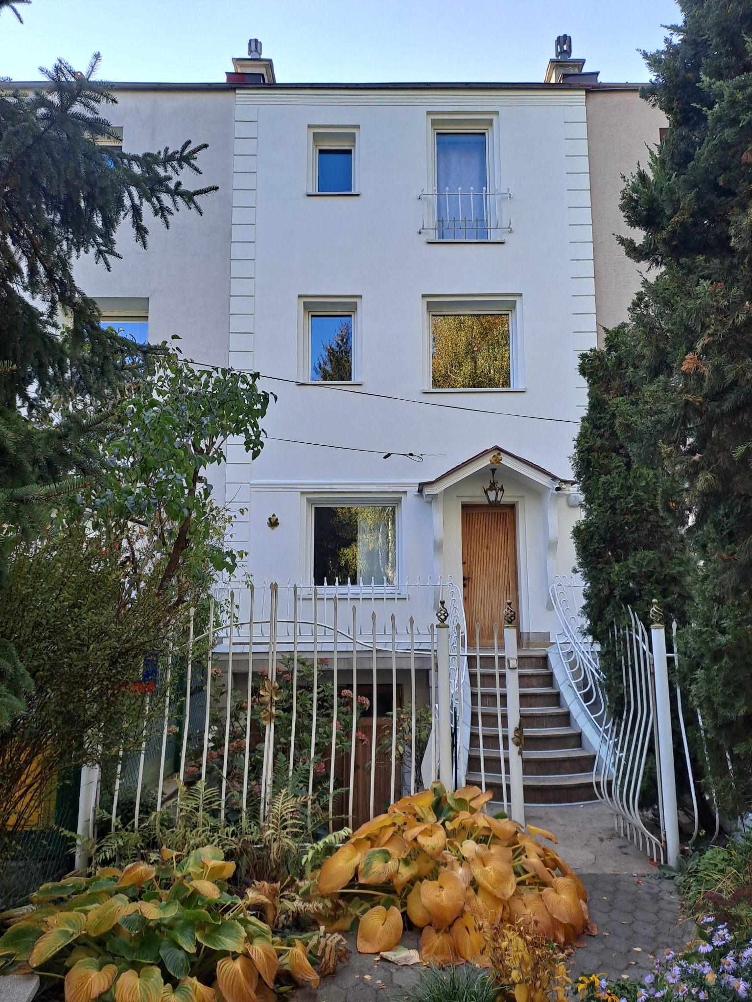 Biała elewacja trzykondygnacyjnego domu z drewnianymi drzwiami wejściowymi, widoczna zza ozdobnego metalowego ogrodzenia i bujnej roślinności.