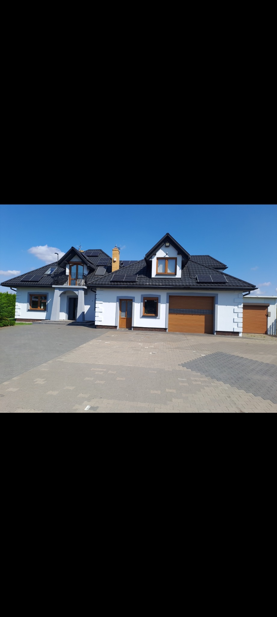 Biały dom jednorodzinny z szarymi akcentami, czarnym dachem i panelami słonecznymi, sfotografowany w słoneczny dzień. Widoczne drewniane okna i drzwi garażowe.