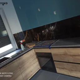 Nowoczesna kuchnia z drewnianymi szafkami, czarnym blatem i marmurowym backsplashiem, niebieskie szafki wiszące, widoczne zabezpieczenia z folii i taśmy malarskiej na blacie, butelka z rozpylaczem...