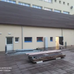 Przygotowania do prac malarskich na zewnątrz budynku: rusztowania, zabezpieczone okna folią, deski i pomalowana elewacja.