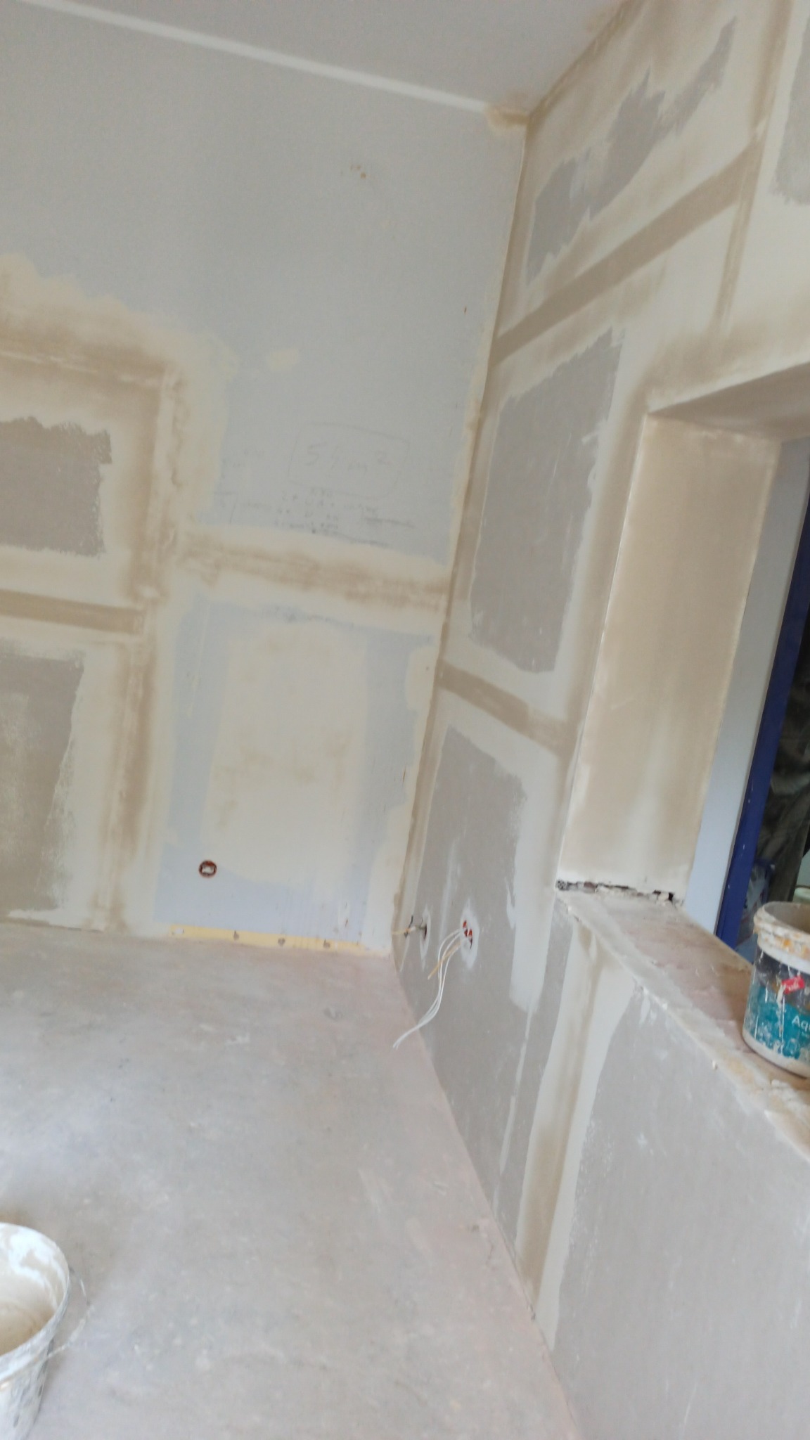 Wnętrze w trakcie szpachlowania ścian, widoczne wystające kable elektryczne, ślady po szpachli na ścianach i podłodze, kubeł z zaprawą malarską w rogu pomieszczenia.