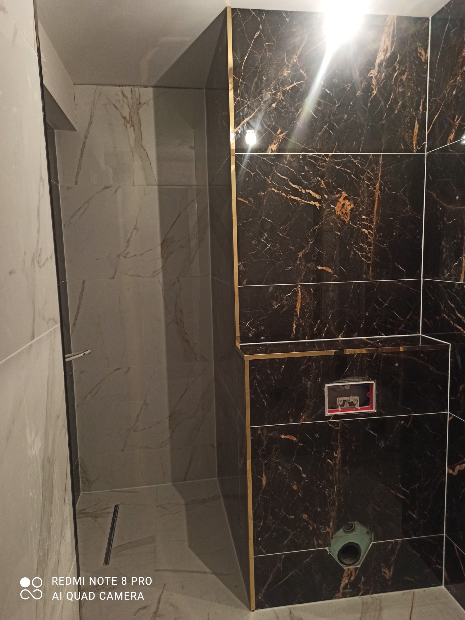 Wykończona łazienka z czarnymi i białymi marmurowymi płytkami, złotymi listwami dekoracyjnymi, odpływem liniowym i przygotowanym miejscem na stelaż podtynkowy WC.