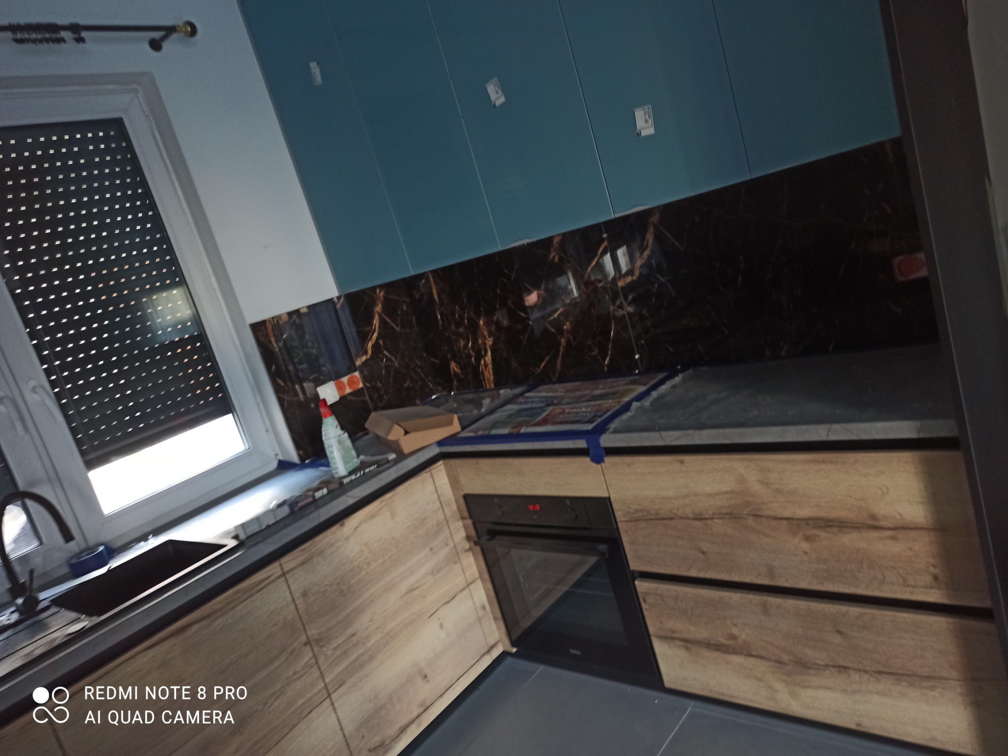 Nowoczesna kuchnia z drewnianymi szafkami, czarnym blatem i marmurowym backsplashiem, niebieskie szafki wiszące, widoczne zabezpieczenia z folii i taśmy malarskiej na blacie, butelka z rozpylaczem...