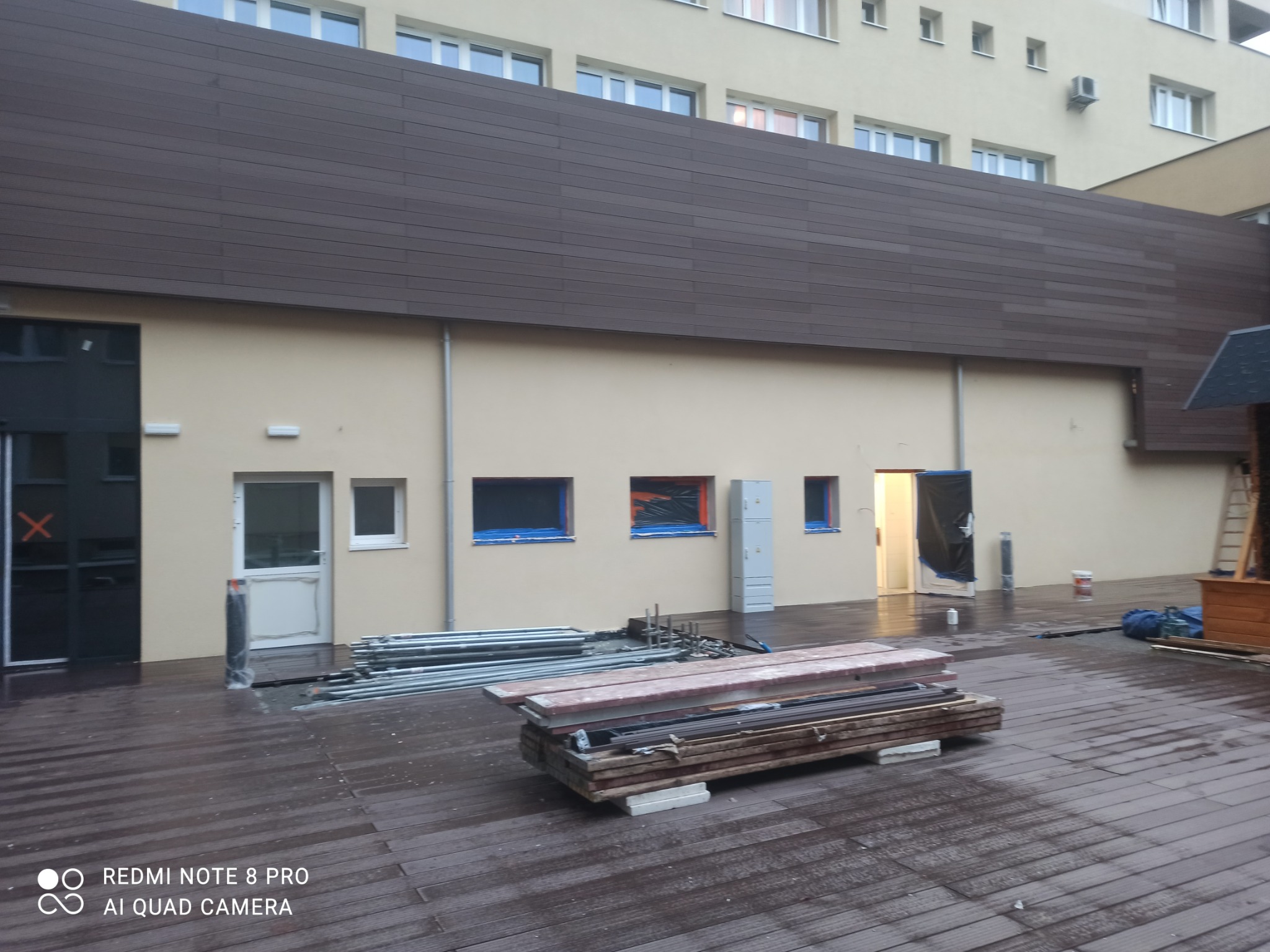 Przygotowania do prac malarskich na zewnątrz budynku: rusztowania, zabezpieczone okna folią, deski i pomalowana elewacja.