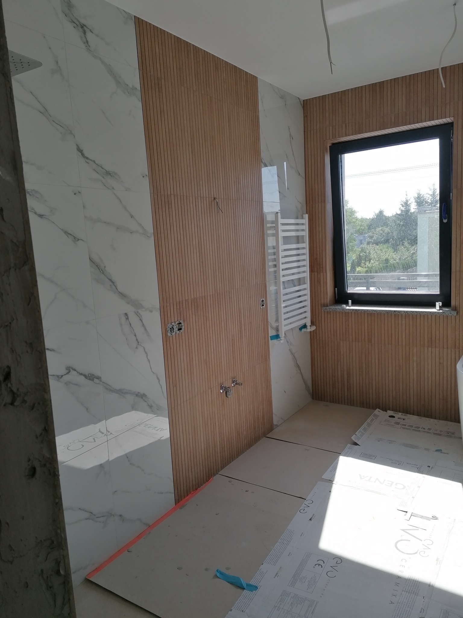 Wnętrze łazienki w trakcie remontu: ściany wyłożone płytkami imitującymi marmur i drewniane panele, widoczne instalacje elektryczne i hydrauliczne, grzejnik drabinkowy, podłoga zabezpieczona kartonem.