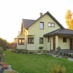 Żółty dom jednorodzinny z ciemnobrązowym dachem i elementami wykończeniowymi, otoczony zadbanym trawnikiem i ozdobnymi rabatami, z widocznym czerwonym samochodem na podjeździe.