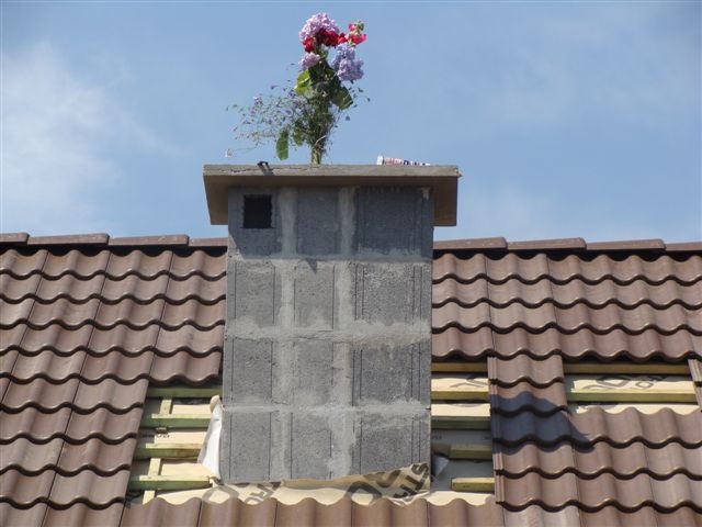 Niedokończony komin z szarego betonu na brązowym dachu z dachówki ceramicznej, ozdobiony bukietem kwiatów na szczycie, widoczne elementy konstrukcji dachu pod dachówką.