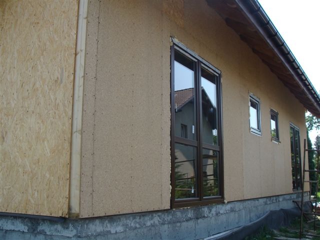 Budowa domu szkieletowego: ściana zewnętrzna z widocznym poszyciem z płyty OSB i fragmentem otynkowanej elewacji, okna z brązowymi ramami.