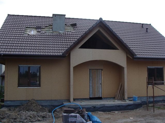 Dom w stanie surowym zamkniętym z brązową dachówką, widoczne uszkodzenia w pokryciu dachowym w okolicy komina, oraz niezabudowany szczyt nad wejściem.