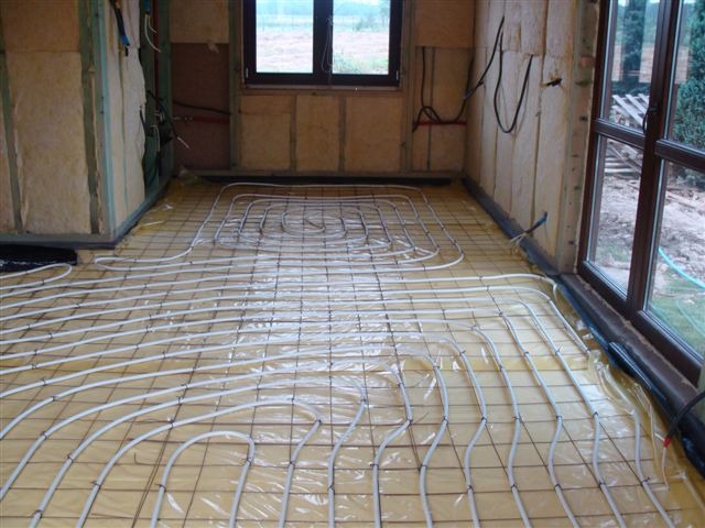 Instalacja ogrzewania podłogowego w domu w trakcie budowy, widoczne rury grzewcze na żółtej folii, ściany z izolacją, okno z widokiem na zewnątrz.