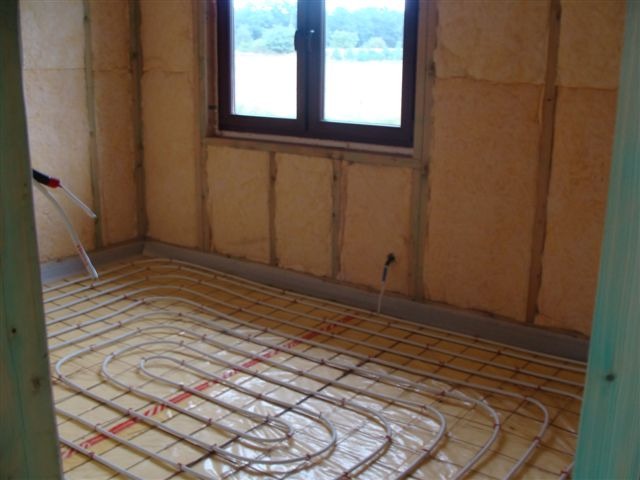 Surowy stan wnętrza domu szkieletowego z widoczną instalacją ogrzewania podłogowego na folii izolacyjnej, ściany wykończone płytami z wełny drzewnej, okno w tle.