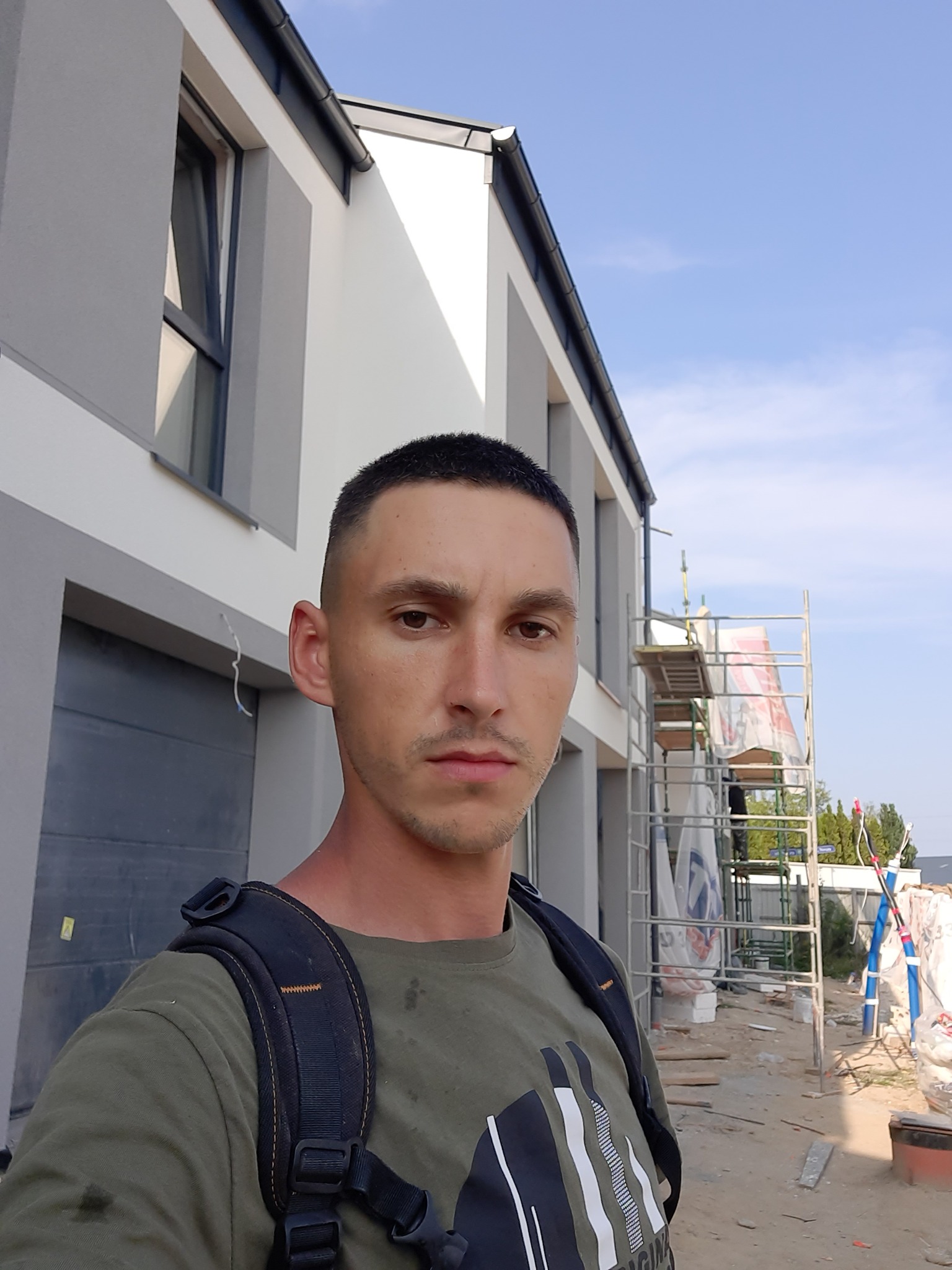 Mężczyzna z plecakiem na tle dwukondygnacyjnego domu w trakcie prac elewacyjnych, widoczne rusztowania.