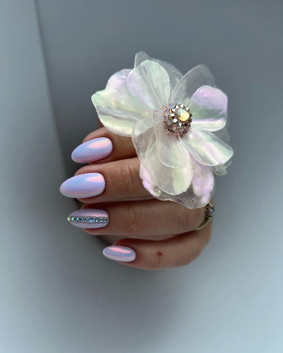 Dłoń z owalnymi paznokciami w kolorze opalizującego fioletu, jeden paznokieć ozdobiony rzędem cyrkonii, dekoracja w kształcie białego kwiatu z opalizującymi płatkami.