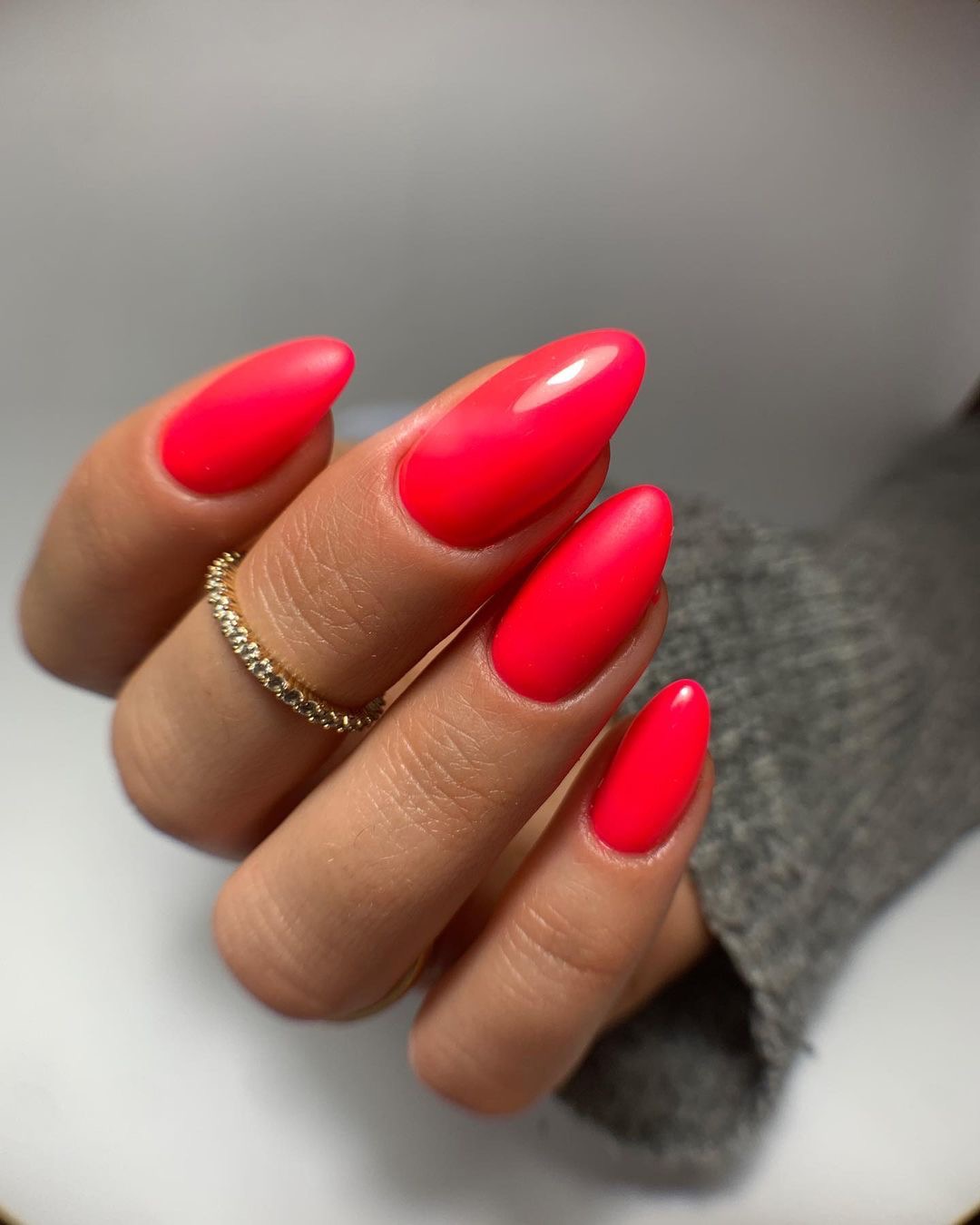 Dłoń z jaskrawoczerwonymi, migdałowymi paznokciami na tle szarego swetra i jasnego tła, ozdobiona pierścionkiem z drobnymi kamieniami na palcu wskazującym.