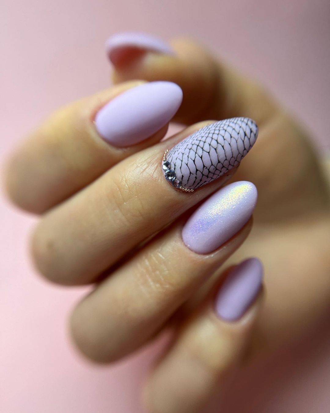 Dłoń z pastelowymi, liliowymi paznokciami; jeden z nich zdobiony czarną siateczką i rzędem małych, srebrnych dżetów przy skórkach.