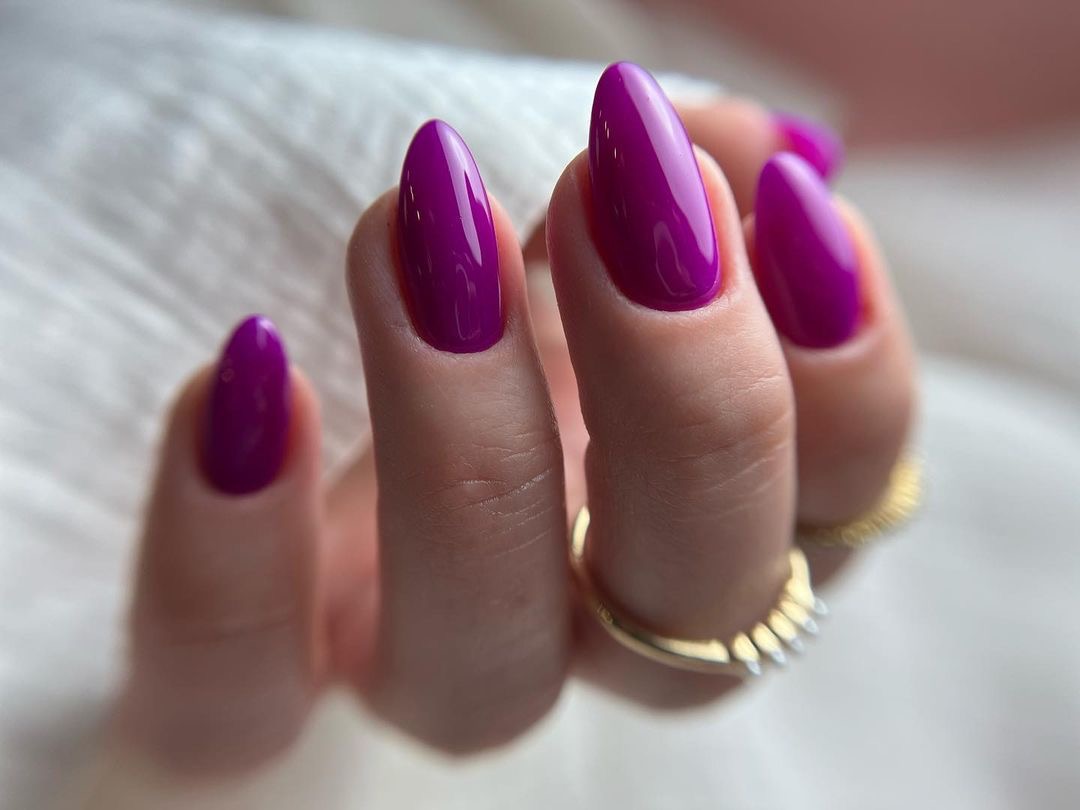 Dłoń z pomalowanymi na intensywny fiolet migdałowymi paznokciami, ozdobiona złotymi pierścionkami na tle białej, miękkiej tkaniny.