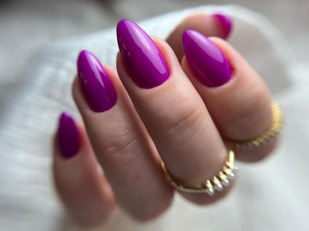 Dłoń z fioletowymi, błyszczącymi paznokciami w kształcie migdała, ozdobiona złotym pierścionkiem z drobnymi perłami na tle białej, miękkiej tkaniny.