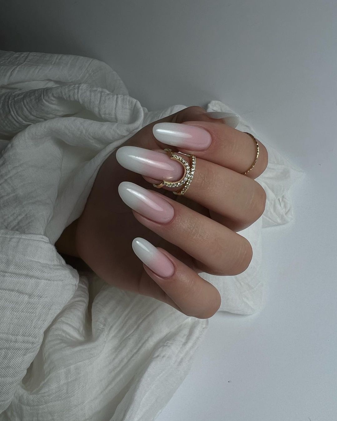 Dłoń z długimi, owalnymi paznokciami w stylu french ombre, ozdobiona dwoma złotymi pierścionkami na palcu serdecznym, na tle białej, pogniecionej tkaniny.