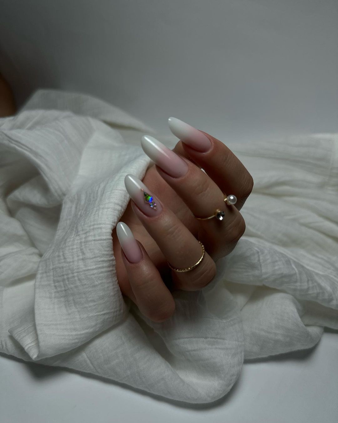 Dłoń z długimi, owalnymi paznokciami w stylu ombre (biały przechodzący w róż) ozdobionymi drobnymi, błyszczącymi kamieniami, na palcach złote pierścionki, ułożona na białej, pogniecionej tkaninie.