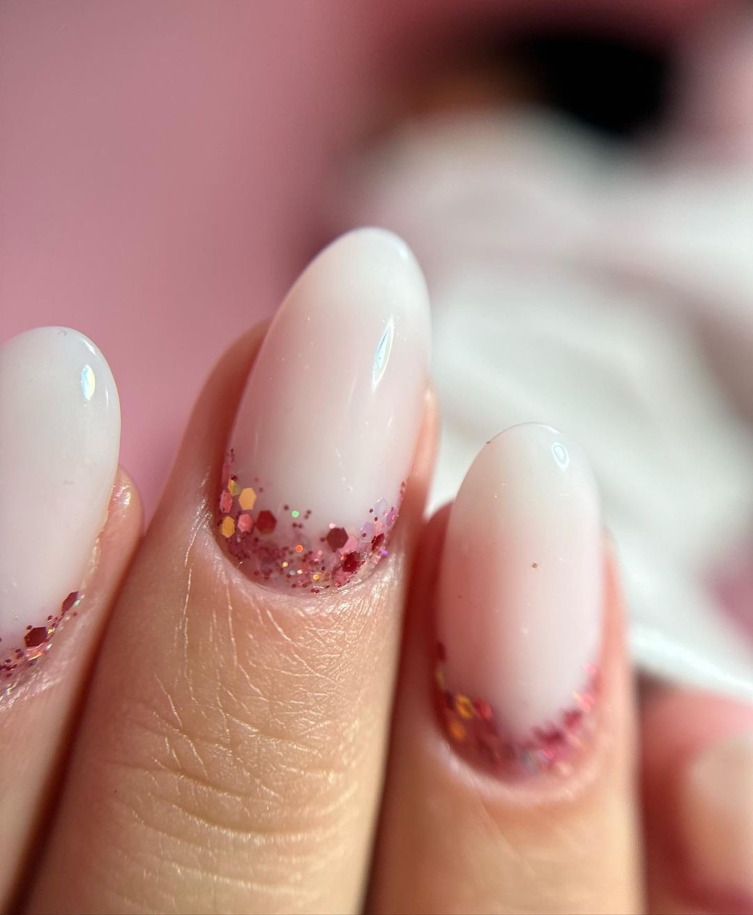 Delikatny manicure french z różowymi brokatowymi końcówkami na migdałowych paznokciach, widoczne drobne zmarszczki na skórze palca.