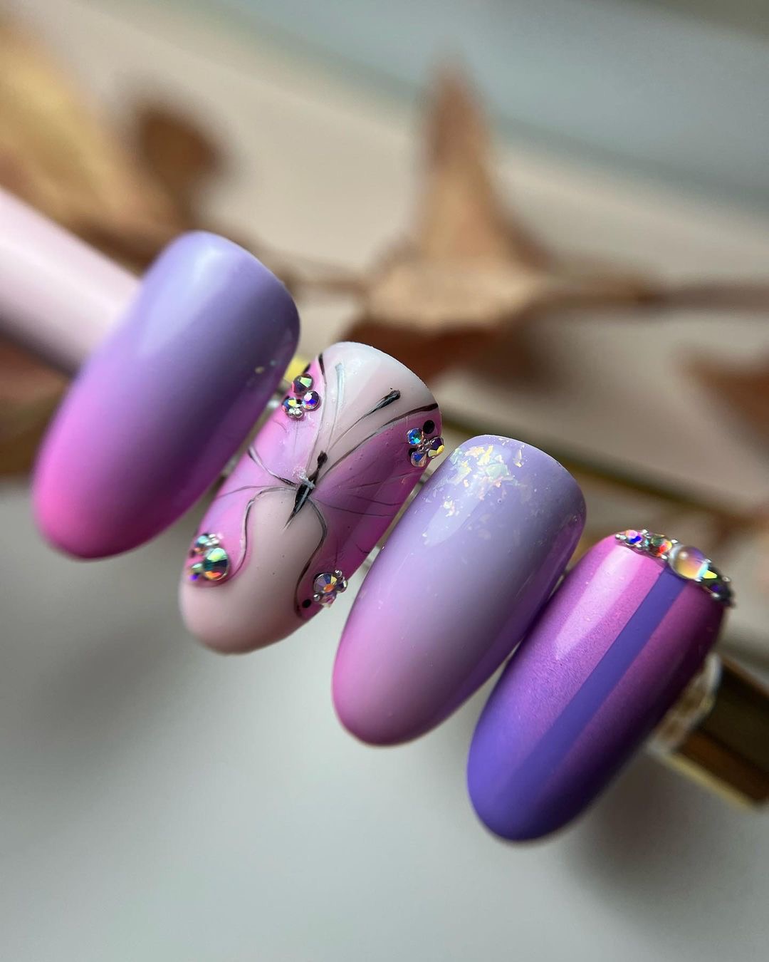 Cztery sztuczne paznokcie w odcieniach fioletu i różu, zdobione motywem motyla i błyszczącymi kamieniami, ustawione na jasnym tle z elementami dekoracyjnymi.